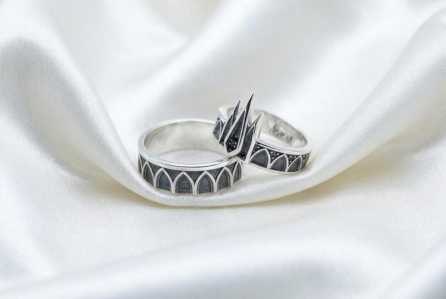 A high-resolution studio photograph of a matching sterling silver gothic wedding ring set resting on a reflective black background. The set features a wide band with a continuous arcade of pointed lancet arches. The companion engagement ring beautifully matches the arch design, forming a dramatic tiara silhouette accented by small, round black gemstones set within the structural peaks. The silver is heavily oxidized.