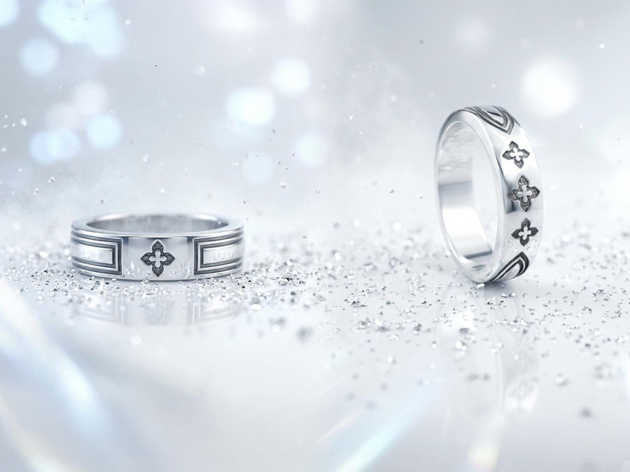 A high-resolution studio shot of a matching sterling silver gothic wedding ring set resting on a sparkling, white surface scattered with tiny crystals. The beautiful set includes a wide men's band and a narrower engagement ring centering a vibrant round red ruby. Both rings feature precisely polished medieval quatrefoil motifs four-lobed geometric shapes set against a deeply oxidized, stippled dark grey background, beautifully highlighting th