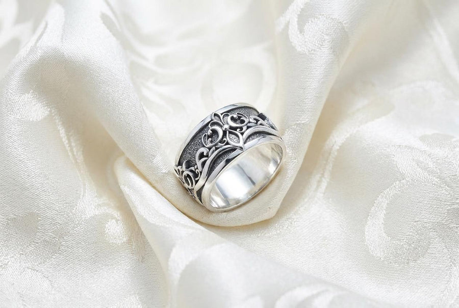 A close-up studio shot of a matching sterling silver gothic wedding ring set resting on a dark reflective surface. The set includes a wide band and a matching V-shaped chevron ring that nests perfectly against it. Both rings feature raised, polished silver scrollwork and fleur-de-lis motifs set against a deeply oxidized, stippled dark grey background. The intricate relief mimics medieval stone carvings, offering a rugged yet highly sophisticated