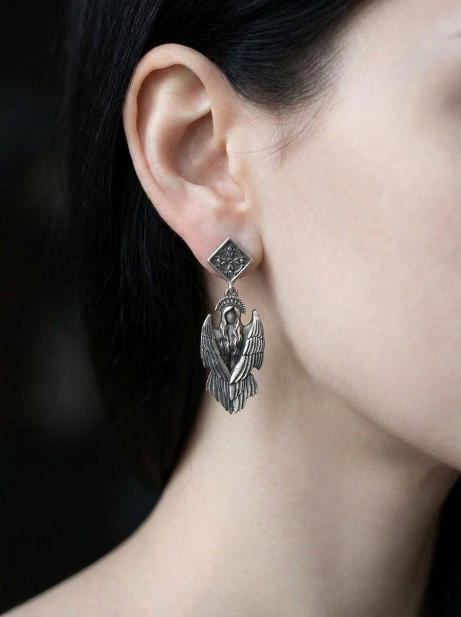 A detailed studio macro shot of a pair of articulated sterling silver earrings on a dark reflective surface. The top stud is a diamond shape enclosing a gothic floral cross. It suspends a highly detailed seraphim angel figure featuring a faceless head crowned with a studded halo. Her large, textured wings fold downwards into a feathered skirt. The silver is oxidized, leaving the recesses dark while the raised feathers gleam. (etsy image id: 8003029872)