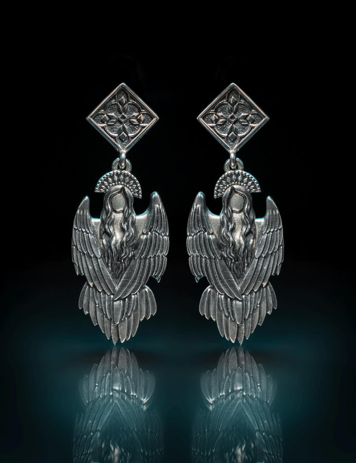 A detailed studio macro shot of a pair of articulated sterling silver earrings on a dark reflective surface. The top stud is a diamond shape enclosing a gothic floral cross. It suspends a highly detailed seraphim angel figure featuring a faceless head crowned with a studded halo. Her large, textured wings fold downwards into a feathered skirt. The silver is oxidized, leaving the recesses dark while the raised feathers gleam. (etsy image id: 8003029872)