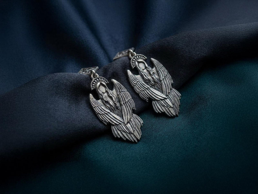 A detailed studio macro shot of a pair of articulated sterling silver earrings on a dark reflective surface. The top stud is a diamond shape enclosing a gothic floral cross. It suspends a highly detailed seraphim angel figure featuring a faceless head crowned with a studded halo. Her large, textured wings fold downwards into a feathered skirt. The silver is oxidized, leaving the recesses dark while the raised feathers gleam. (etsy image id: 8003029872)