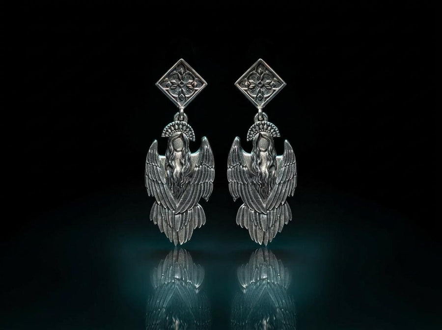 A detailed studio macro shot of a pair of articulated sterling silver earrings on a dark reflective surface. The top stud is a diamond shape enclosing a gothic floral cross. It suspends a highly detailed seraphim angel figure featuring a faceless head crowned with a studded halo. Her large, textured wings fold downwards into a feathered skirt. The silver is oxidized, leaving the recesses dark while the raised feathers gleam. (etsy image id: 8003029872)