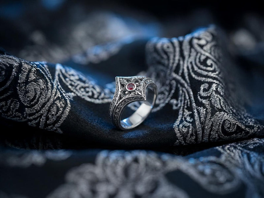 A close-up studio photograph of a matching sterling silver gothic wedding ring set resting on a highly reflective black surface. The set includes a wide band and a pointed, V-shaped chevron ring. Both bands feature deeply carved, raised leaf and vine scrollwork. The engagement ring centers a round, dark red garnet gemstone. The silver is heavily oxidized, creating a stark contrast between the bright ridges and the dark recesses.