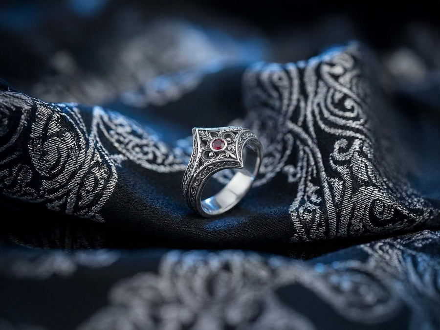 A close-up studio photograph of a matching sterling silver gothic wedding ring set resting on a highly reflective black surface. The set includes a wide band and a pointed, V-shaped chevron ring. Both bands feature deeply carved, raised leaf and vine scrollwork. The engagement ring centers a round, dark red garnet gemstone. The silver is heavily oxidized, creating a stark contrast between the bright ridges and the dark recesses.