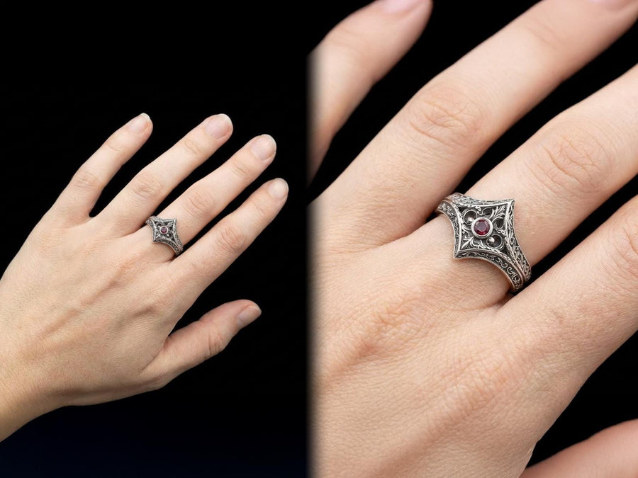 A close-up studio photograph of a matching sterling silver gothic wedding ring set resting on a highly reflective black surface. The set includes a wide band and a pointed, V-shaped chevron ring. Both bands feature deeply carved, raised leaf and vine scrollwork. The engagement ring centers a round, dark red garnet gemstone. The silver is heavily oxidized, creating a stark contrast between the bright ridges and the dark recesses.