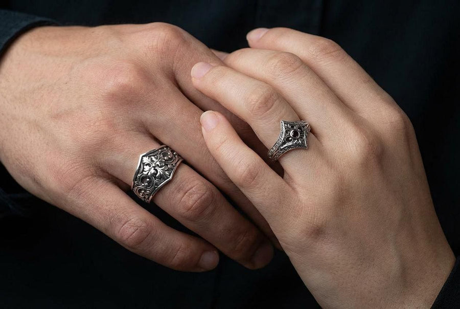 A close-up studio photograph of a matching sterling silver gothic wedding ring set resting on a highly reflective black surface. The set includes a wide band and a pointed, V-shaped chevron ring. Both bands feature deeply carved, raised leaf and vine scrollwork. The engagement ring centers a round, dark red garnet gemstone. The silver is heavily oxidized, creating a stark contrast between the bright ridges and the dark recesses.