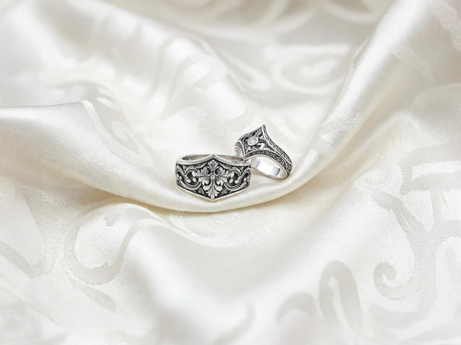 A close-up studio photograph of a matching sterling silver gothic wedding ring set resting on a highly reflective black surface. The set includes a wide band and a pointed, V-shaped chevron ring. Both bands feature deeply carved, raised leaf and vine scrollwork. The engagement ring centers a round, dark red garnet gemstone. The silver is heavily oxidized, creating a stark contrast between the bright ridges and the dark recesses.