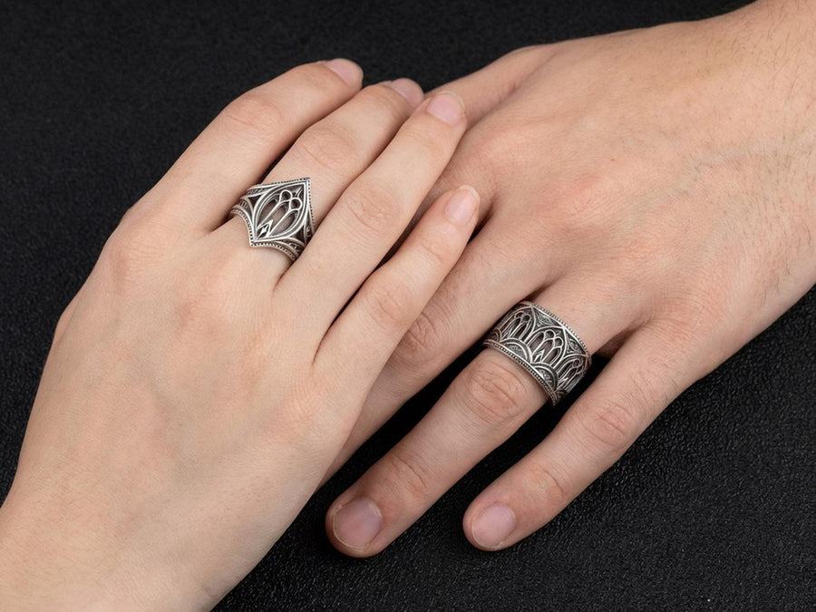 A high-resolution macro shot of a wide sterling silver wedding band resting on a sparkling, icy white surface. The ring features an intricate openwork design mimicking a Gothic cathedral window, complete with pointed lancet arches and delicate stone tracery. The polished silver highlights contrast beautifully against the dark, oxidized background, emphasizing the architectural depth of the piece.