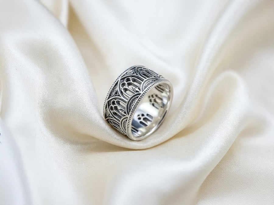 A high-resolution macro shot of a wide sterling silver wedding band resting on a sparkling, icy white surface. The ring features an intricate openwork design mimicking a Gothic cathedral window, complete with pointed lancet arches and delicate stone tracery. The polished silver highlights contrast beautifully against the dark, oxidized background, emphasizing the architectural depth of the piece.