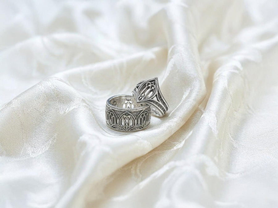 A high-resolution macro shot of a wide sterling silver wedding band resting on a sparkling, icy white surface. The ring features an intricate openwork design mimicking a Gothic cathedral window, complete with pointed lancet arches and delicate stone tracery. The polished silver highlights contrast beautifully against the dark, oxidized background, emphasizing the architectural depth of the piece.