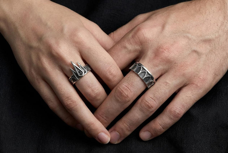 A high-resolution studio photograph of a matching sterling silver gothic wedding ring set resting on a reflective black background. The set features a wide band with a continuous arcade of pointed lancet arches. The companion engagement ring beautifully matches the arch design, forming a dramatic tiara silhouette accented by small, round black gemstones set within the structural peaks. The silver is heavily oxidized.