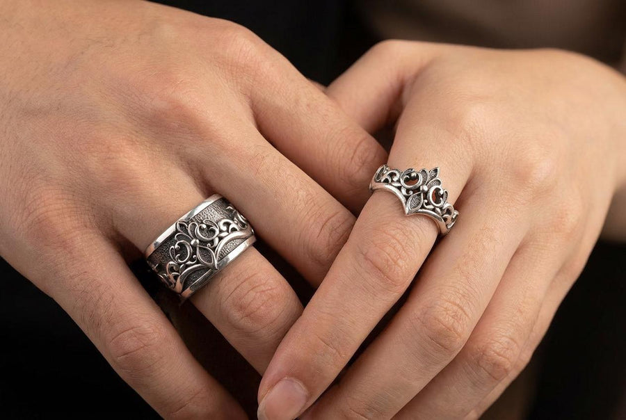 A close-up studio shot of a matching sterling silver gothic wedding ring set resting on a dark reflective surface. The set includes a wide band and a matching V-shaped chevron ring that nests perfectly against it. Both rings feature raised, polished silver scrollwork and fleur-de-lis motifs set against a deeply oxidized, stippled dark grey background. The intricate relief mimics medieval stone carvings, offering a rugged yet highly sophisticated