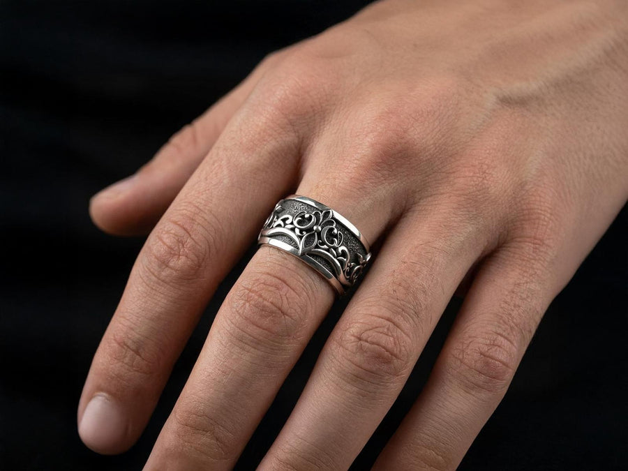 A close-up studio shot of a matching sterling silver gothic wedding ring set resting on a dark reflective surface. The set includes a wide band and a matching V-shaped chevron ring that nests perfectly against it. Both rings feature raised, polished silver scrollwork and fleur-de-lis motifs set against a deeply oxidized, stippled dark grey background. The intricate relief mimics medieval stone carvings, offering a rugged yet highly sophisticated