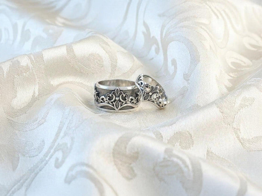A close-up studio shot of a matching sterling silver gothic wedding ring set resting on a dark reflective surface. The set includes a wide band and a matching V-shaped chevron ring that nests perfectly against it. Both rings feature raised, polished silver scrollwork and fleur-de-lis motifs set against a deeply oxidized, stippled dark grey background. The intricate relief mimics medieval stone carvings, offering a rugged yet highly sophisticated