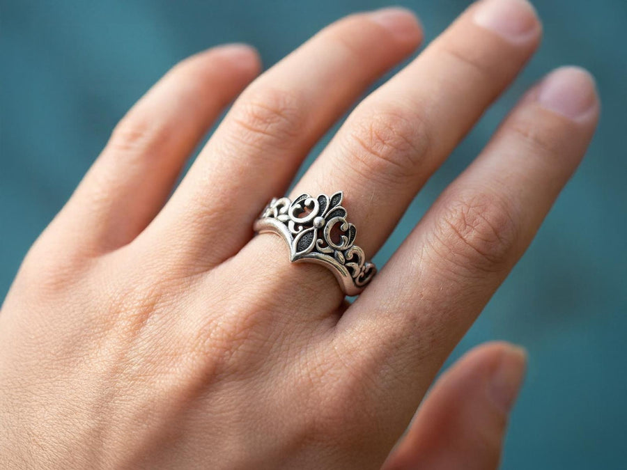 A close-up studio shot of a matching sterling silver gothic wedding ring set resting on a dark reflective surface. The set includes a wide band and a matching V-shaped chevron ring that nests perfectly against it. Both rings feature raised, polished silver scrollwork and fleur-de-lis motifs set against a deeply oxidized, stippled dark grey background. The intricate relief mimics medieval stone carvings, offering a rugged yet highly sophisticated