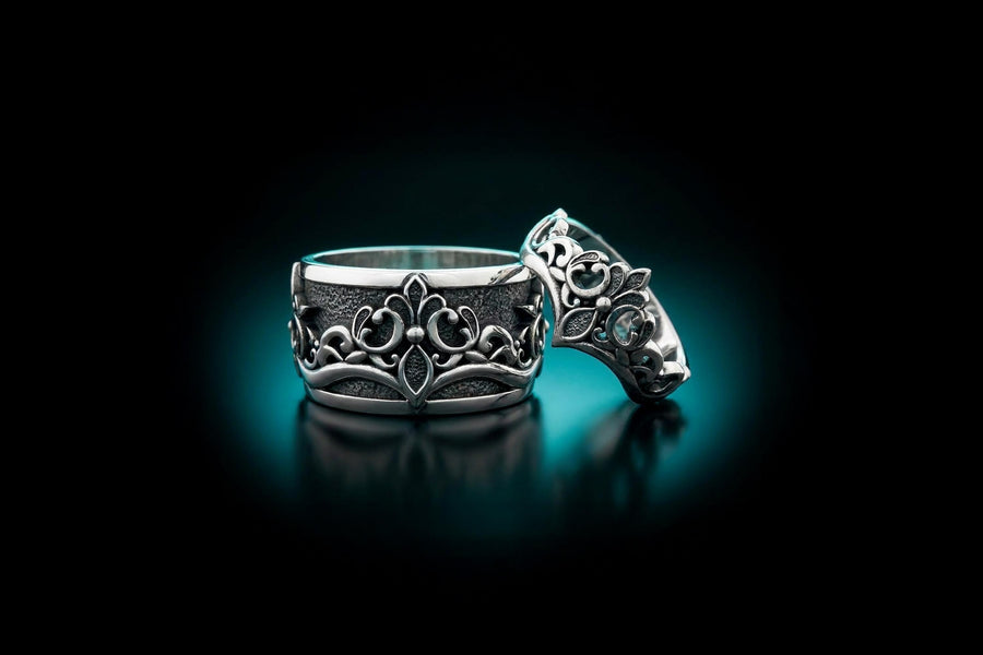 A close-up studio shot of a matching sterling silver gothic wedding ring set resting on a dark reflective surface. The set includes a wide band and a matching V-shaped chevron ring that nests perfectly against it. Both rings feature raised, polished silver scrollwork and fleur-de-lis motifs set against a deeply oxidized, stippled dark grey background. The intricate relief mimics medieval stone carvings, offering a rugged yet highly sophisticated