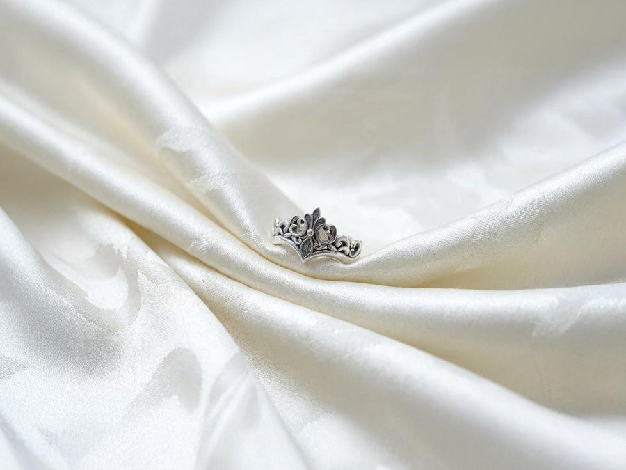 A close-up studio shot of a matching sterling silver gothic wedding ring set resting on a dark reflective surface. The set includes a wide band and a matching V-shaped chevron ring that nests perfectly against it. Both rings feature raised, polished silver scrollwork and fleur-de-lis motifs set against a deeply oxidized, stippled dark grey background. The intricate relief mimics medieval stone carvings, offering a rugged yet highly sophisticated