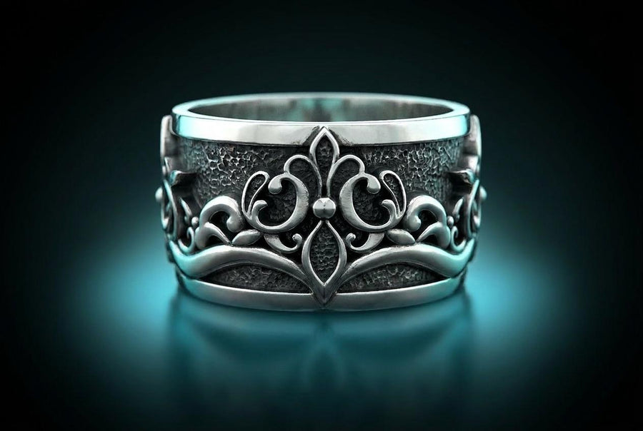 A close-up studio shot of a matching sterling silver gothic wedding ring set resting on a dark reflective surface. The set includes a wide band and a matching V-shaped chevron ring that nests perfectly against it. Both rings feature raised, polished silver scrollwork and fleur-de-lis motifs set against a deeply oxidized, stippled dark grey background. The intricate relief mimics medieval stone carvings, offering a rugged yet highly sophisticated