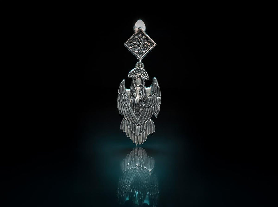 A detailed studio macro shot of a pair of articulated sterling silver earrings on a dark reflective surface. The top stud is a diamond shape enclosing a gothic floral cross. It suspends a highly detailed seraphim angel figure featuring a faceless head crowned with a studded halo. Her large, textured wings fold downwards into a feathered skirt. The silver is oxidized, leaving the recesses dark while the raised feathers gleam. (etsy image id: 8003029872)