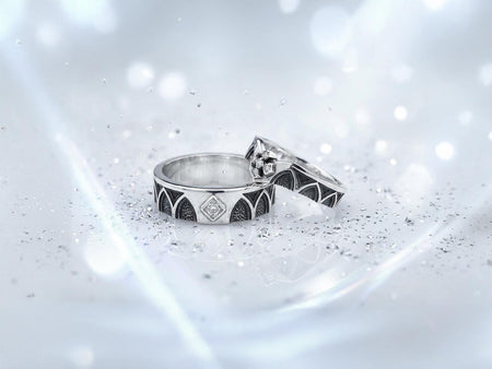 A high-resolution macro shot of a matching sterling silver gothic wedding ring set resting on a dark reflective surface. The set includes a wide men's band with pointed lancet arches and a chevron engagement ring with a white diamond cross cluster. Both feature deeply oxidized, stippled backgrounds that highlight the polished silver tracery and the sparkling square-cut center diamond.