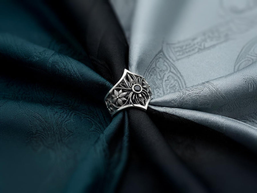 A close-up studio photograph of a highly detailed sterling silver gothic ring resting on a pitch-black reflective surface. The focal point is an intricate three-dimensional spider, featuring a round, bezel-set black onyx gemstone as its abdomen. The spider is positioned at the center of a pointed gothic shield resembling a rose window or web. The side bands are adorned with deeply carved, repeating geometric rosettes.