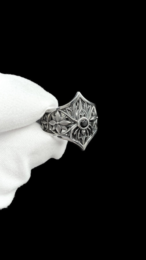 A close-up studio photograph of a highly detailed sterling silver gothic ring resting on a pitch-black reflective surface. The focal point is an intricate three-dimensional spider, featuring a round, bezel-set black onyx gemstone as its abdomen. The spider is positioned at the center of a pointed gothic shield resembling a rose window or web. The side bands are adorned with deeply carved, repeating geometric rosettes.