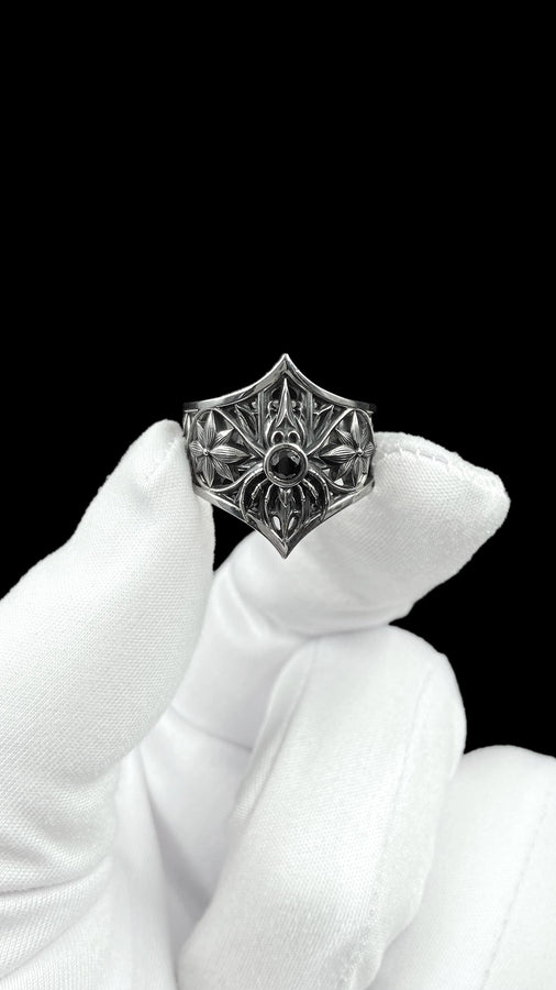A close-up studio photograph of a highly detailed sterling silver gothic ring resting on a pitch-black reflective surface. The focal point is an intricate three-dimensional spider, featuring a round, bezel-set black onyx gemstone as its abdomen. The spider is positioned at the center of a pointed gothic shield resembling a rose window or web. The side bands are adorned with deeply carved, repeating geometric rosettes.