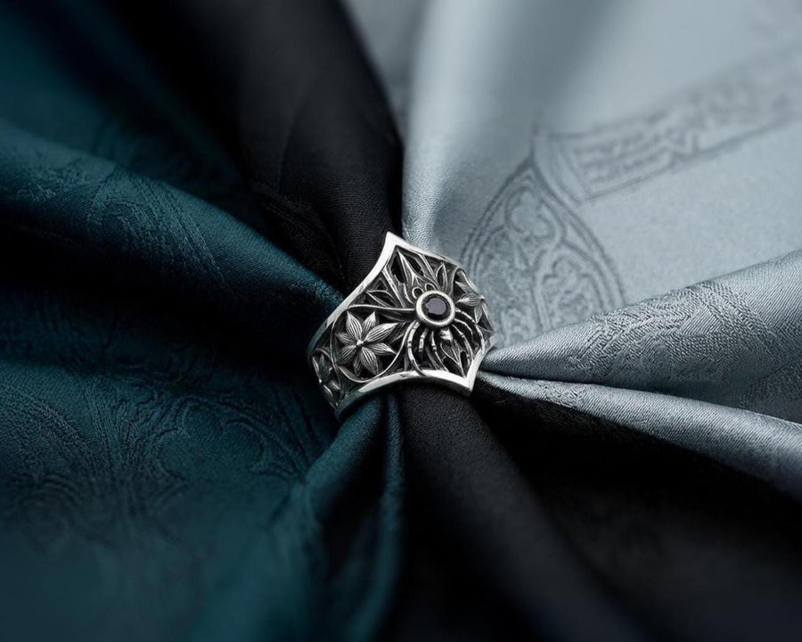 A close-up studio photograph of a highly detailed sterling silver gothic ring resting on a pitch-black reflective surface. The focal point is an intricate three-dimensional spider, featuring a round, bezel-set black onyx gemstone as its abdomen. The spider is positioned at the center of a pointed gothic shield resembling a rose window or web. The side bands are adorned with deeply carved, repeating geometric rosettes.
