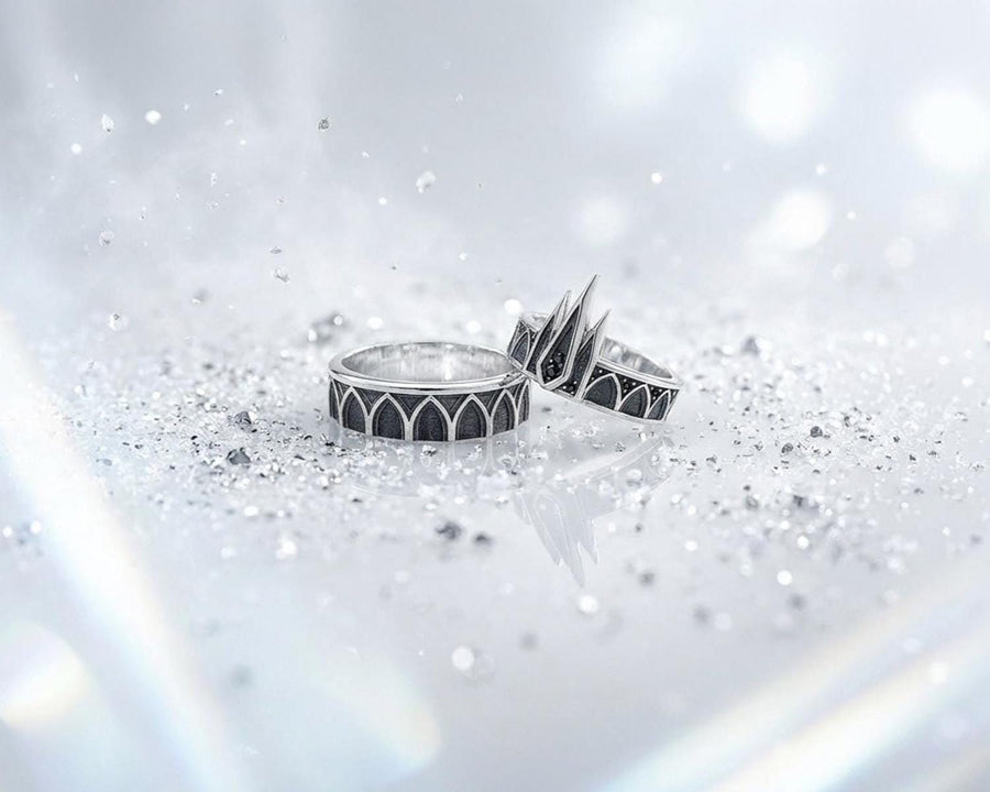 A high-resolution studio photograph of a matching sterling silver gothic wedding ring set resting on a reflective black background. The set features a wide band with a continuous arcade of pointed lancet arches. The companion engagement ring beautifully matches the arch design, forming a dramatic tiara silhouette accented by small, round black gemstones set within the structural peaks. The silver is heavily oxidized.