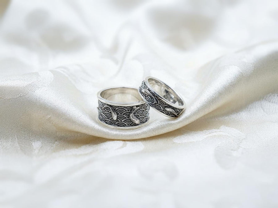 A close-up studio photograph of a matching pair of 925 sterling silver wedding bands resting on a bright, reflective surface scattered with water droplets or ice crystals. The rings feature a deeply engraved, continuous pattern of traditional Japanese Seigaiha ocean waves. Overlaid on the wave texture are sculpted, three-dimensional koi fish swimming elegantly along the curved bands. The recessed areas of the silver are deeply oxidized to a dark