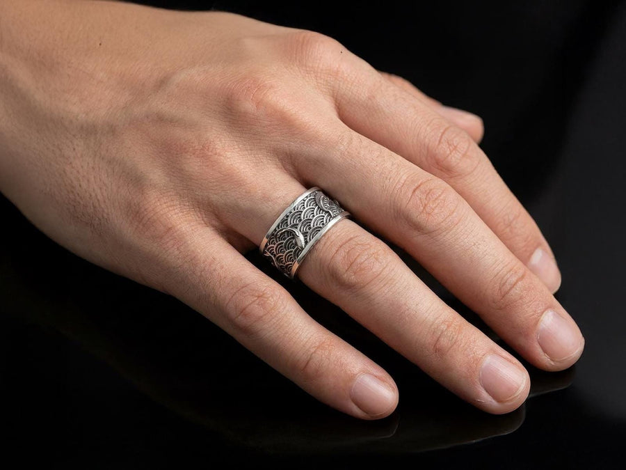 A close-up studio photograph of a matching pair of 925 sterling silver wedding bands resting on a bright, reflective surface scattered with water droplets or ice crystals. The rings feature a deeply engraved, continuous pattern of traditional Japanese Seigaiha ocean waves. Overlaid on the wave texture are sculpted, three-dimensional koi fish swimming elegantly along the curved bands. The recessed areas of the silver are deeply oxidized to a dark