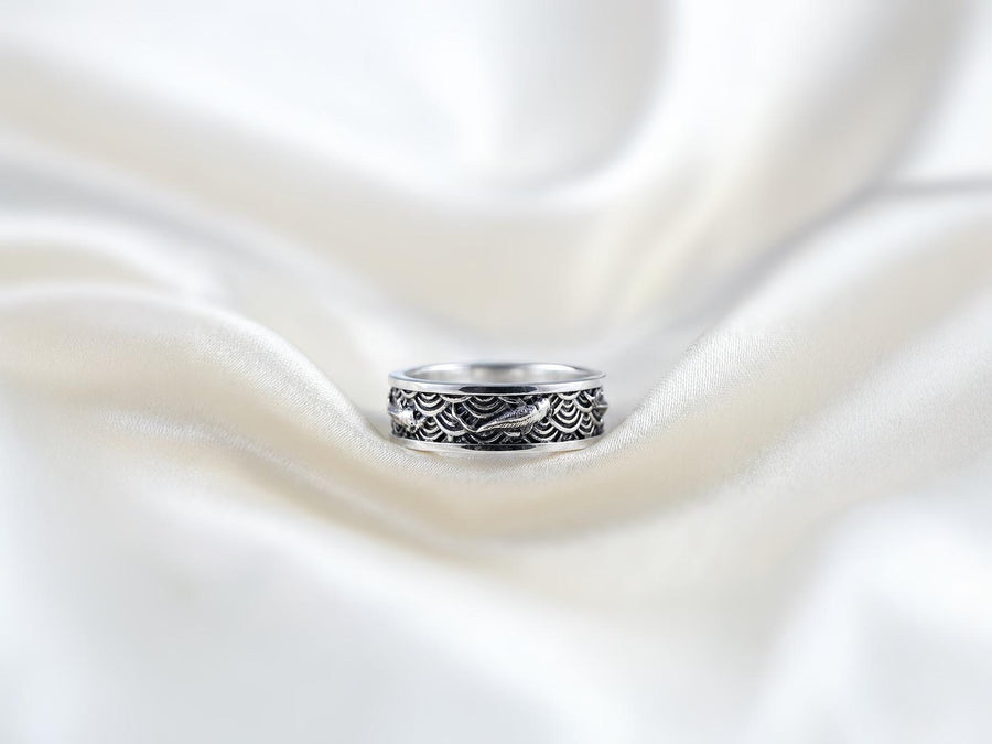 A close-up studio photograph of a matching pair of 925 sterling silver wedding bands resting on a bright, reflective surface scattered with water droplets or ice crystals. The rings feature a deeply engraved, continuous pattern of traditional Japanese Seigaiha ocean waves. Overlaid on the wave texture are sculpted, three-dimensional koi fish swimming elegantly along the curved bands. The recessed areas of the silver are deeply oxidized to a dark