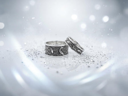 A close-up studio photograph of a matching pair of 925 sterling silver wedding bands resting on a bright, reflective surface scattered with water droplets or ice crystals. The rings feature a deeply engraved, continuous pattern of traditional Japanese Seigaiha ocean waves. Overlaid on the wave texture are sculpted, three-dimensional koi fish swimming elegantly along the curved bands. The recessed areas of the silver are deeply oxidized to a dark