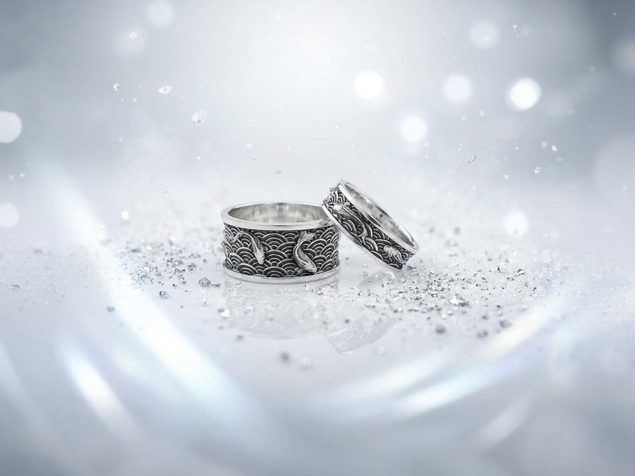 A close-up studio photograph of a matching pair of 925 sterling silver wedding bands resting on a bright, reflective surface scattered with water droplets or ice crystals. The rings feature a deeply engraved, continuous pattern of traditional Japanese Seigaiha ocean waves. Overlaid on the wave texture are sculpted, three-dimensional koi fish swimming elegantly along the curved bands. The recessed areas of the silver are deeply oxidized to a dark