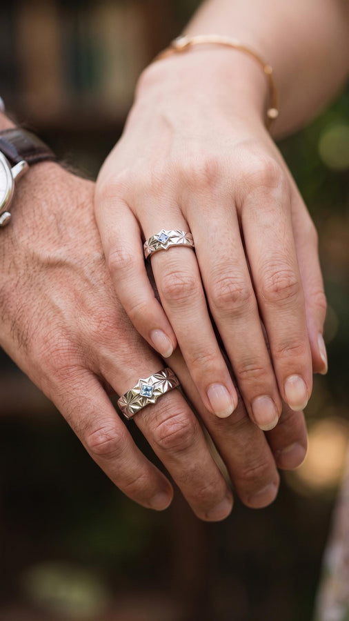 A close-up studio photograph of a matching pair of 925 sterling silver wedding bands resting on a bright, reflective white surface scattered with tiny crystals. The rings feature a continuous, deeply engraved pattern of geometric starbursts or celestial stars with radiating lines. The primary band has a square, princess-cut light blue gemstone, likely blue topaz or aquamarine, bezel-set directly into the center of the prominent front-facing star.