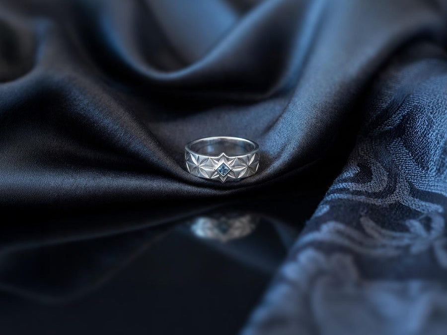 A close-up studio photograph of a matching pair of 925 sterling silver wedding bands resting on a bright, reflective white surface scattered with tiny crystals. The rings feature a continuous, deeply engraved pattern of geometric starbursts or celestial stars with radiating lines. The primary band has a square, princess-cut light blue gemstone, likely blue topaz or aquamarine, bezel-set directly into the center of the prominent front-facing star.