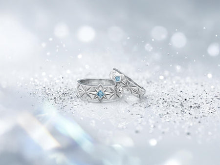 A close-up studio photograph of a matching pair of 925 sterling silver wedding bands resting on a bright, reflective white surface scattered with tiny crystals. The rings feature a continuous, deeply engraved pattern of geometric starbursts or celestial stars with radiating lines. The primary band has a square, princess-cut light blue gemstone, likely blue topaz or aquamarine, bezel-set directly into the center of the prominent front-facing star.