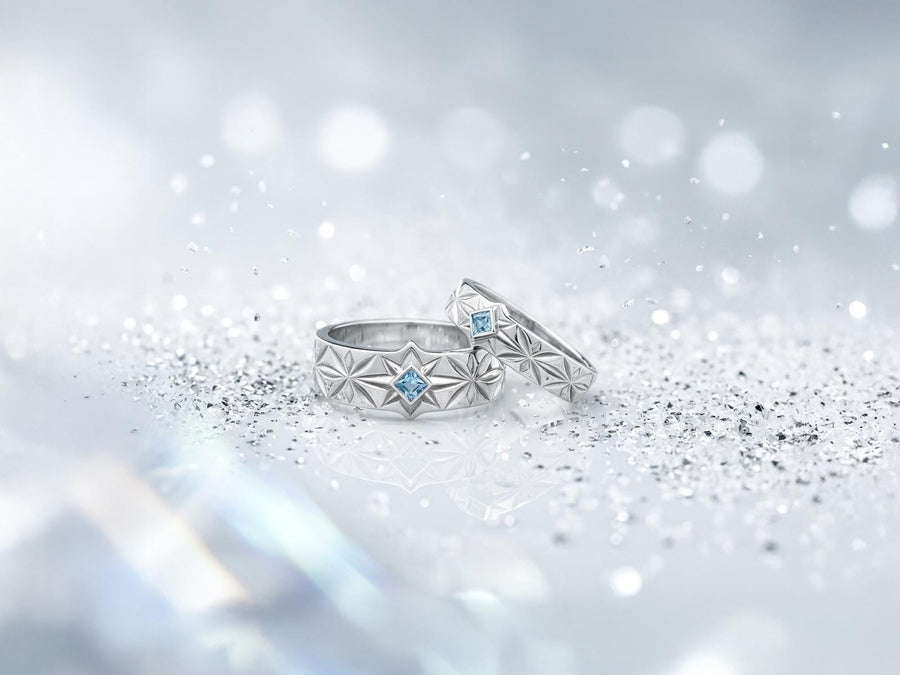 A close-up studio photograph of a matching pair of 925 sterling silver wedding bands resting on a bright, reflective white surface scattered with tiny crystals. The rings feature a continuous, deeply engraved pattern of geometric starbursts or celestial stars with radiating lines. The primary band has a square, princess-cut light blue gemstone, likely blue topaz or aquamarine, bezel-set directly into the center of the prominent front-facing star.