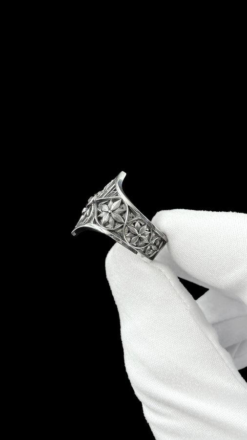 A close-up studio photograph of a highly detailed sterling silver gothic ring resting on a pitch-black reflective surface. The focal point is an intricate three-dimensional spider, featuring a round, bezel-set black onyx gemstone as its abdomen. The spider is positioned at the center of a pointed gothic shield resembling a rose window or web. The side bands are adorned with deeply carved, repeating geometric rosettes.