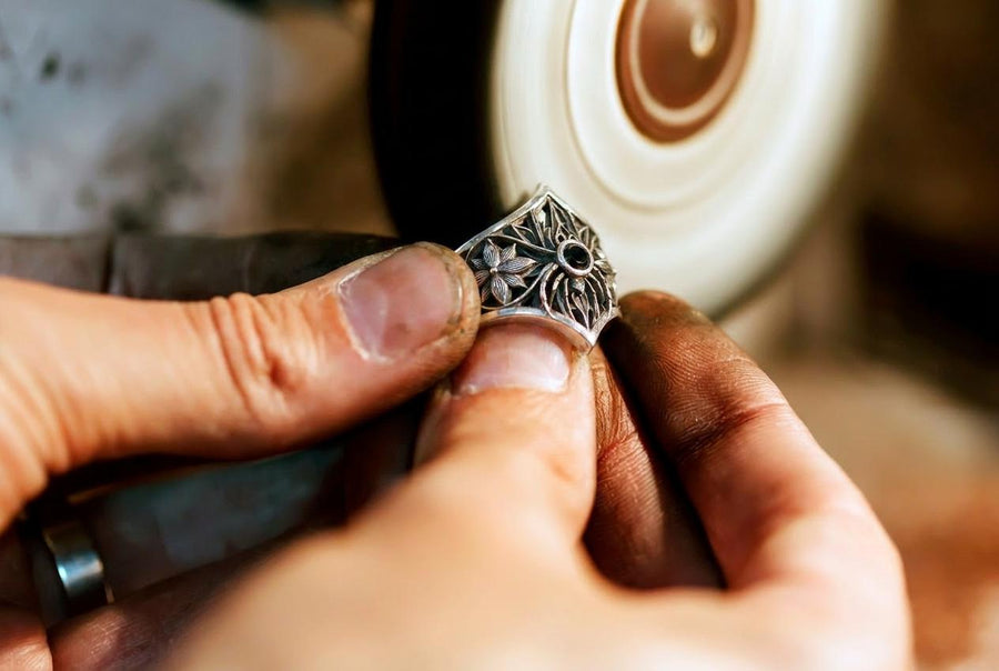 A close-up studio photograph of a highly detailed sterling silver gothic ring resting on a pitch-black reflective surface. The focal point is an intricate three-dimensional spider, featuring a round, bezel-set black onyx gemstone as its abdomen. The spider is positioned at the center of a pointed gothic shield resembling a rose window or web. The side bands are adorned with deeply carved, repeating geometric rosettes.