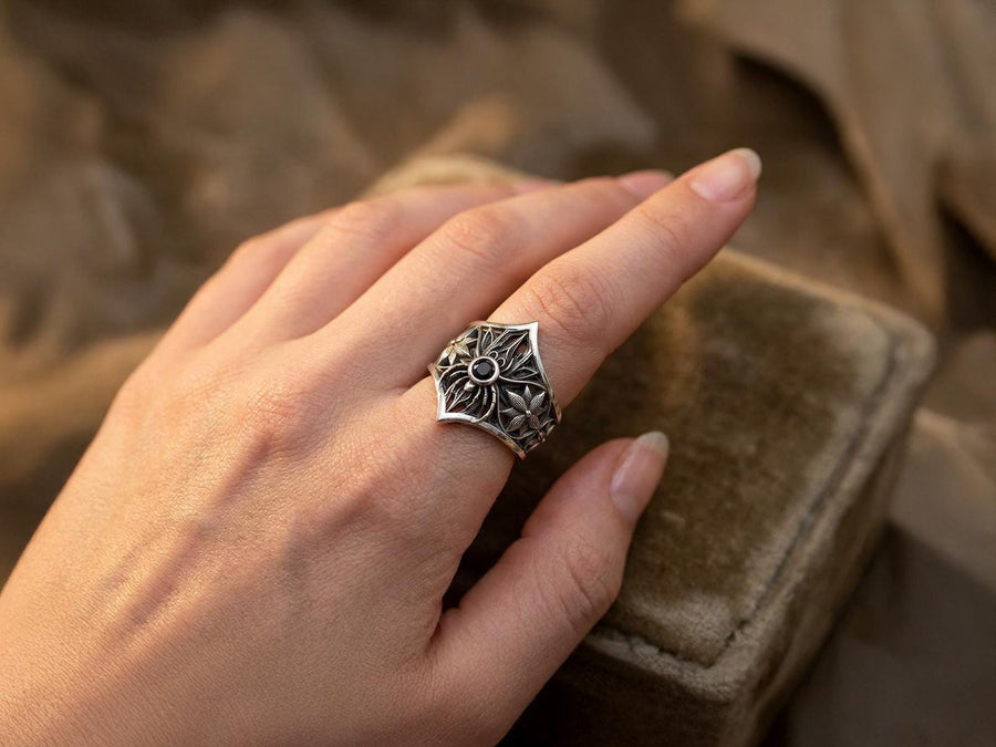 A close-up studio photograph of a highly detailed sterling silver gothic ring resting on a pitch-black reflective surface. The focal point is an intricate three-dimensional spider, featuring a round, bezel-set black onyx gemstone as its abdomen. The spider is positioned at the center of a pointed gothic shield resembling a rose window or web. The side bands are adorned with deeply carved, repeating geometric rosettes.