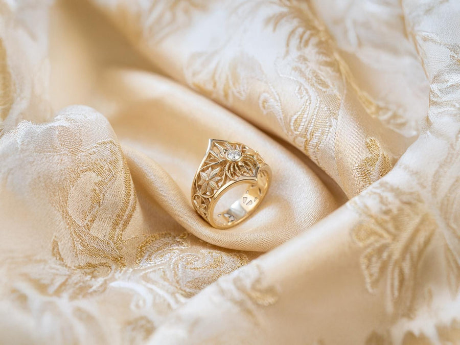A close-up studio photograph of a highly detailed sterling silver gothic ring resting on a pitch-black reflective surface. The focal point is an intricate three-dimensional spider, featuring a round, bezel-set black onyx gemstone as its abdomen. The spider is positioned at the center of a pointed gothic shield resembling a rose window or web. The side bands are adorned with deeply carved, repeating geometric rosettes.