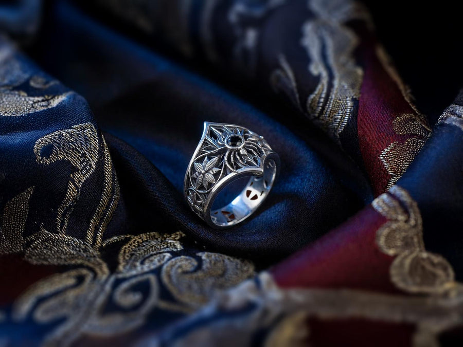 A close-up studio photograph of a highly detailed sterling silver gothic ring resting on a pitch-black reflective surface. The focal point is an intricate three-dimensional spider, featuring a round, bezel-set black onyx gemstone as its abdomen. The spider is positioned at the center of a pointed gothic shield resembling a rose window or web. The side bands are adorned with deeply carved, repeating geometric rosettes.