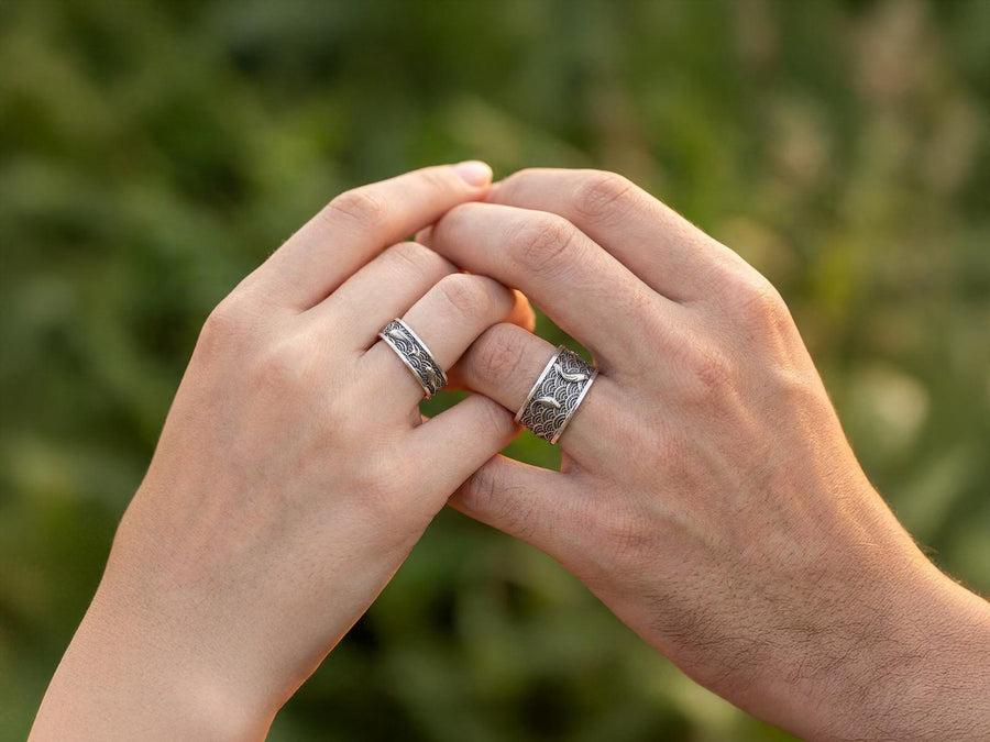 A close-up studio photograph of a matching pair of 925 sterling silver wedding bands resting on a bright, reflective surface scattered with water droplets or ice crystals. The rings feature a deeply engraved, continuous pattern of traditional Japanese Seigaiha ocean waves. Overlaid on the wave texture are sculpted, three-dimensional koi fish swimming elegantly along the curved bands. The recessed areas of the silver are deeply oxidized to a dark