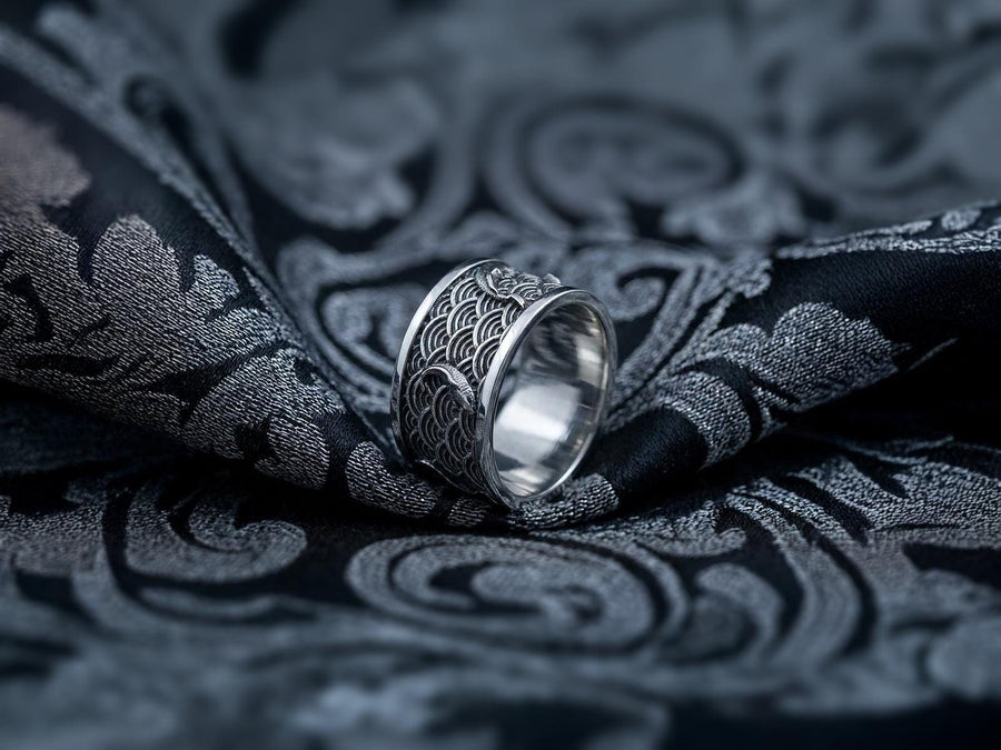 A close-up studio photograph of a matching pair of 925 sterling silver wedding bands resting on a bright, reflective surface scattered with water droplets or ice crystals. The rings feature a deeply engraved, continuous pattern of traditional Japanese Seigaiha ocean waves. Overlaid on the wave texture are sculpted, three-dimensional koi fish swimming elegantly along the curved bands. The recessed areas of the silver are deeply oxidized to a dark
