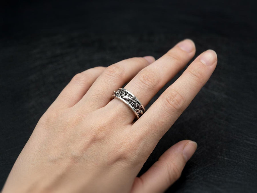 A close-up studio photograph of a matching pair of 925 sterling silver wedding bands resting on a bright, reflective surface scattered with water droplets or ice crystals. The rings feature a deeply engraved, continuous pattern of traditional Japanese Seigaiha ocean waves. Overlaid on the wave texture are sculpted, three-dimensional koi fish swimming elegantly along the curved bands. The recessed areas of the silver are deeply oxidized to a dark