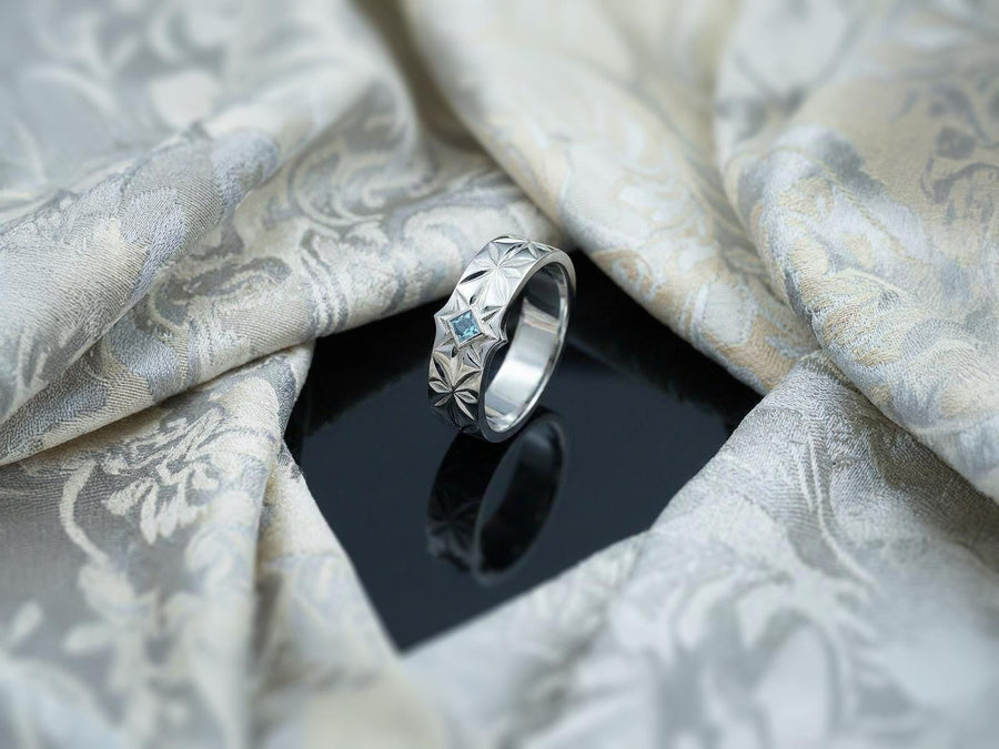 A close-up studio photograph of a matching pair of 925 sterling silver wedding bands resting on a bright, reflective white surface scattered with tiny crystals. The rings feature a continuous, deeply engraved pattern of geometric starbursts or celestial stars with radiating lines. The primary band has a square, princess-cut light blue gemstone, likely blue topaz or aquamarine, bezel-set directly into the center of the prominent front-facing star.
