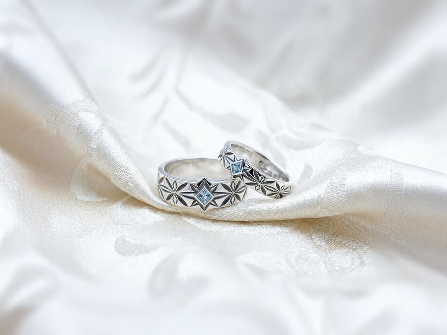 A close-up studio photograph of a matching pair of 925 sterling silver wedding bands resting on a bright, reflective white surface scattered with tiny crystals. The rings feature a continuous, deeply engraved pattern of geometric starbursts or celestial stars with radiating lines. The primary band has a square, princess-cut light blue gemstone, likely blue topaz or aquamarine, bezel-set directly into the center of the prominent front-facing star.