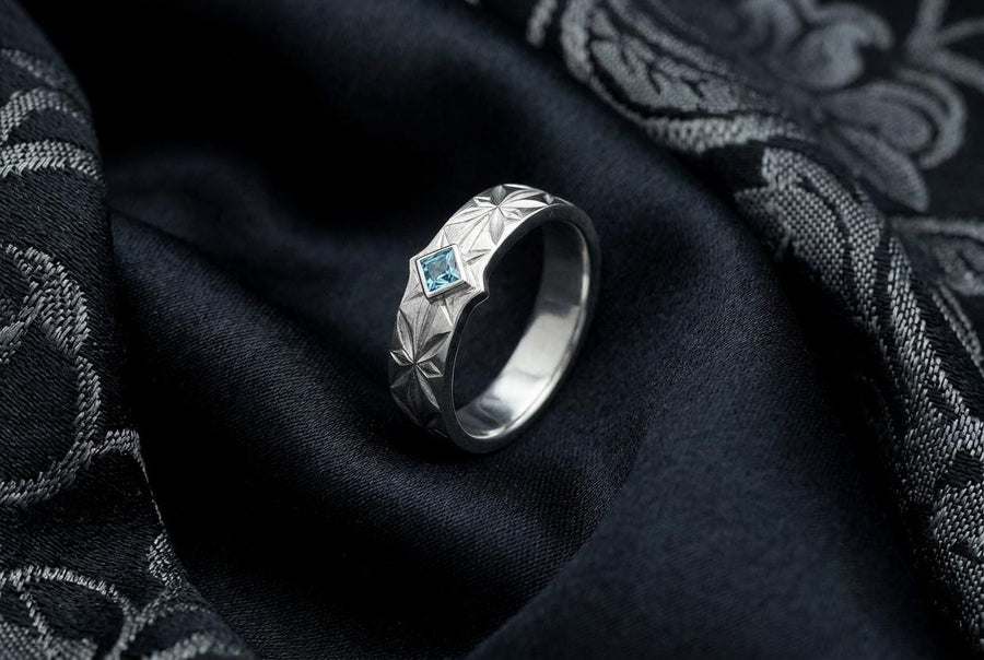 A close-up studio photograph of a matching pair of 925 sterling silver wedding bands resting on a bright, reflective white surface scattered with tiny crystals. The rings feature a continuous, deeply engraved pattern of geometric starbursts or celestial stars with radiating lines. The primary band has a square, princess-cut light blue gemstone, likely blue topaz or aquamarine, bezel-set directly into the center of the prominent front-facing star.